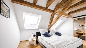 Une chambre avec un plafond en pente, des poutres en bois, une lucarne, un lit avec une literie blanche et des oreillers bleus, une table de chevet avec une lampe et une petite étagère à côté du lit.