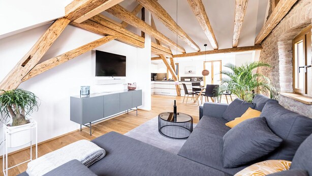 Un loft moderne avec des poutres apparentes, un canapé gris, un téléviseur et une cuisine en arrière-plan.