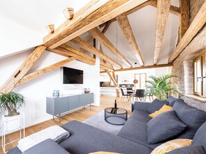 Ein modernes Loft mit freiliegenden Holzbalken, einem grauen Sofa, einem Fernseher und einer Küche im Hintergrund.