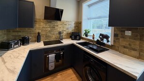 Une cuisine moderne avec des meubles de couleur sombre, une cuisinière noire, un évier et un lave-vaisselle. Il y a une fenêtre avec des stores et une petite plante sur le rebord de la fenêtre.