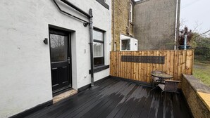 Une terrasse en bois avec une table et des chaises en verre, une porte noire et un mur blanc avec une fenêtre.