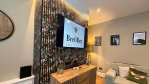 Un salon moderne avec un téléviseur fixé au mur, qui est décoré d’un motif floral, une console en bois agrémentée d’objets décoratifs, un canapé gris avec un coussin vert et des œuvres d’art encadrées accrochées au mur.