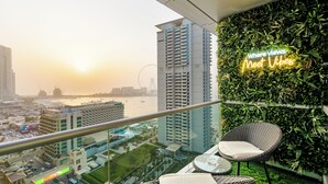 A balcony with wicker chairs, a neon sign, a view of a cityscape, and a building with a green wall. - Grand JBR PentHouse Style (Dubai)