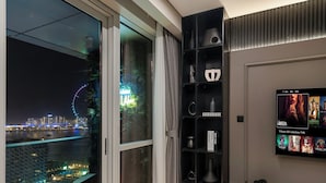 A modern living room with a large TV, a sliding glass door leading to a balcony with a city view, and a dark wood shelving unit. - Grand JBR PentHouse Style (Dubai)