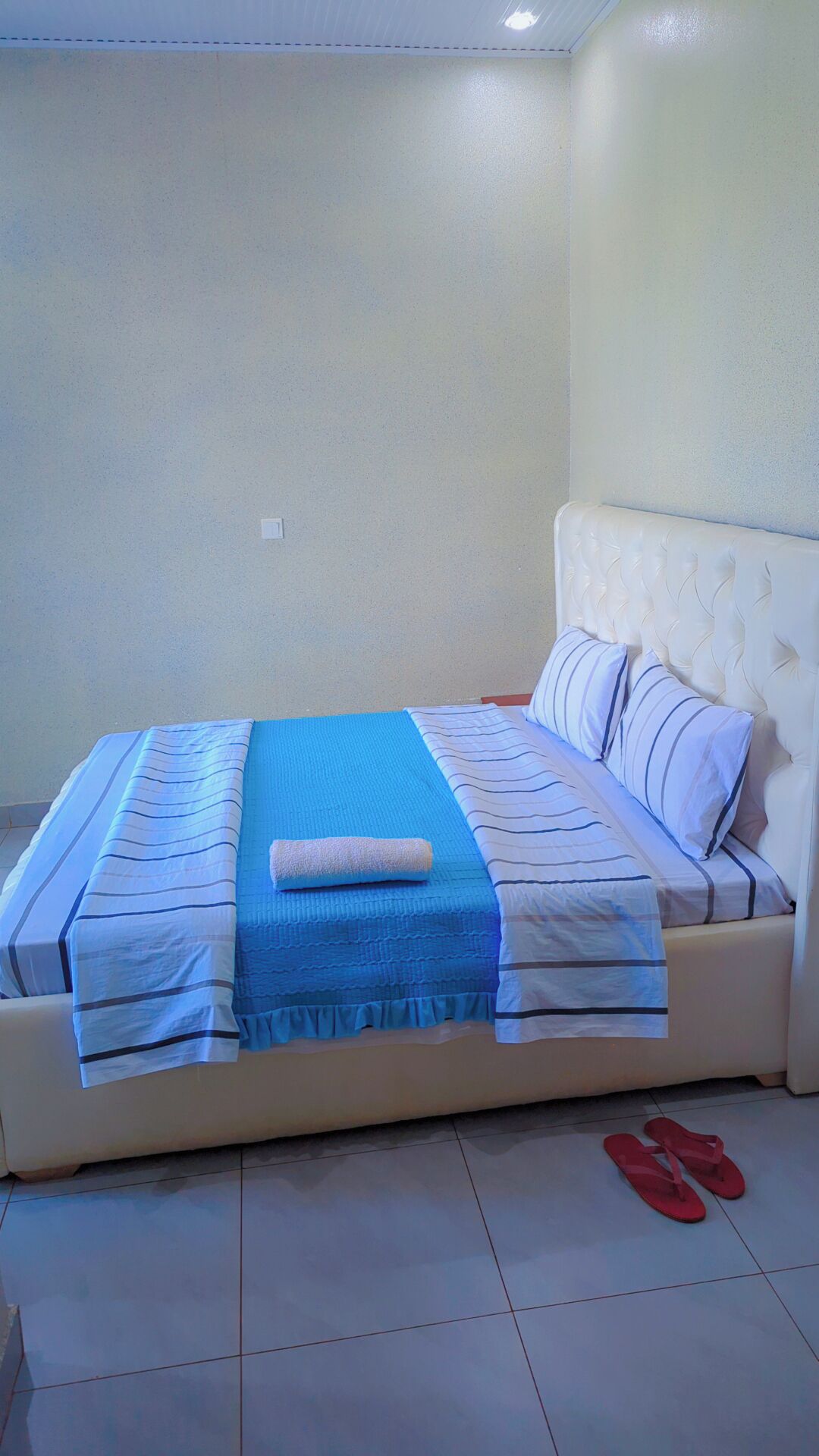 A neatly made bed with a blue blanket and striped pillows, a white headboard, and red slippers on the floor.