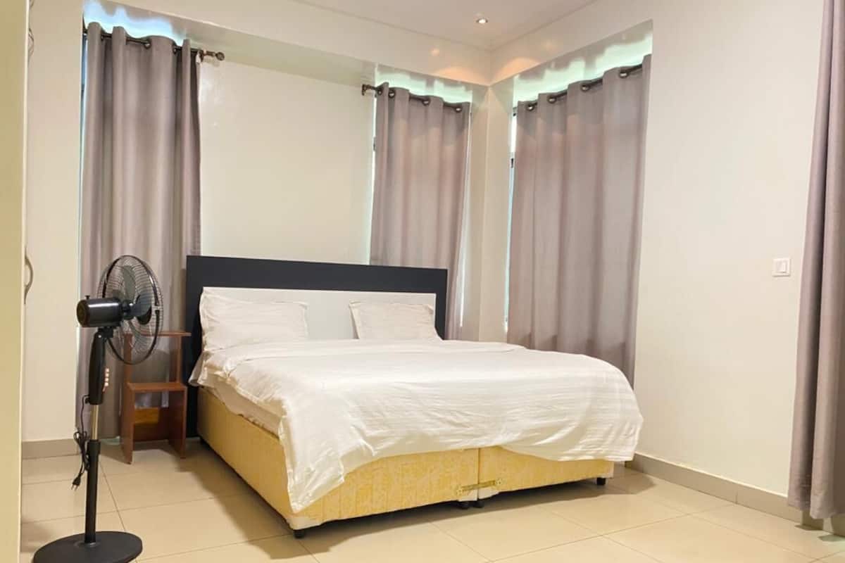 A bedroom with a bed, curtains, a standing fan, and a small wooden table.