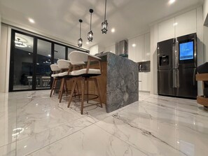A modern kitchen with a marble countertop, bar stools, a refrigerator, and pendant lighting.