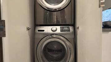 Un appareil combiné lave-linge et sèche-linge à chargement frontal, compact et en acier inoxydable, installé dans un meuble blanc, avec un plan de travail en marbre en dessous.