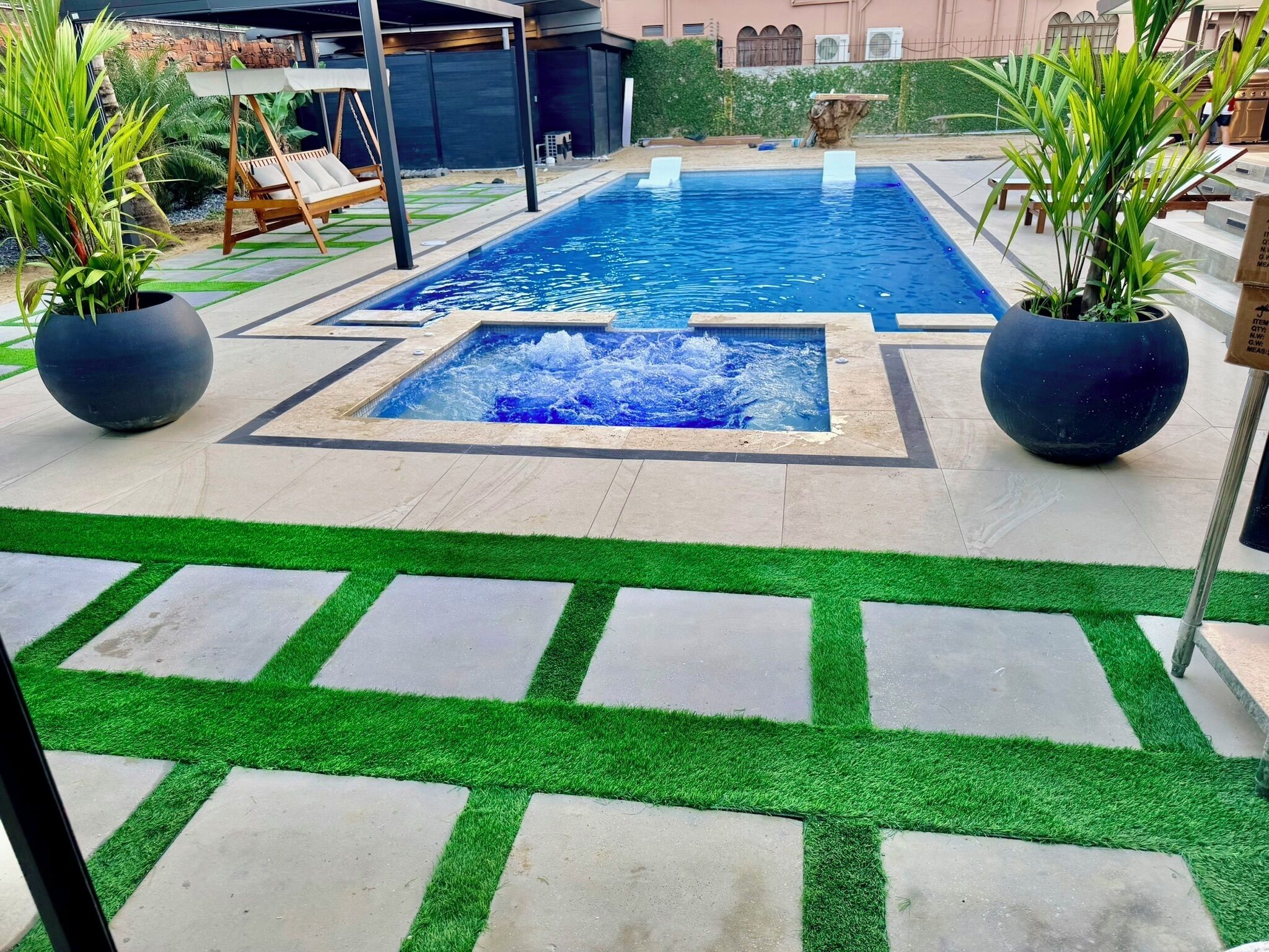 Una piscina rectangular con un elemento acuático central, rodeada por un patio con césped artificial y plantas en macetas.
