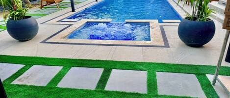 Una piscina rectangular con un elemento acuático central, rodeada por un patio con césped artificial y plantas en macetas.
