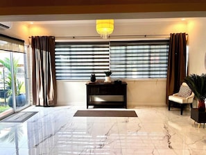 Una habitación amplia con piso de mármol, una consola negra, una silla con cojín estampado y un ventanal con persianas.