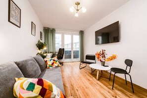 Ein modernes Wohnzimmer mit einem grauen Sofa, einem bunten Kissen, einem kleinen runden Tisch mit Stühlen und einem großen Fenster mit Vorhängen.