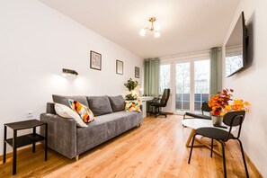 Un salon moderne avec un canapé gris, une petite table d’appoint, une table à manger avec deux chaises et une grande fenêtre avec des rideaux.