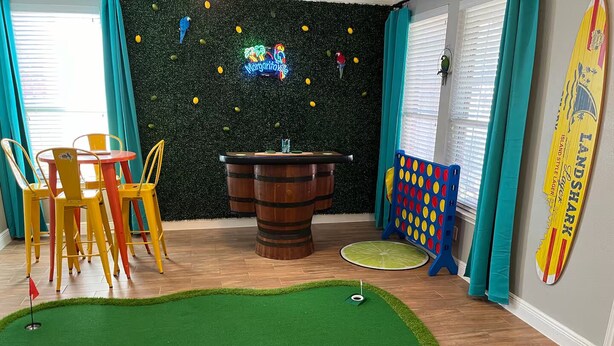 Une pièce avec un tapis de gazon synthétique vert, un tabouret de bar en bois, un jeu de fléchettes et des chaises colorées.