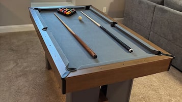 Ein Billardtisch mit Kugeln und Spielstöcken, ein Fenster mit Jalousien und ein graues Sofa in einem Raum mit Teppichboden.