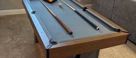 A pool table with balls and cues, a window with blinds, and a grey couch in a room with carpet flooring.