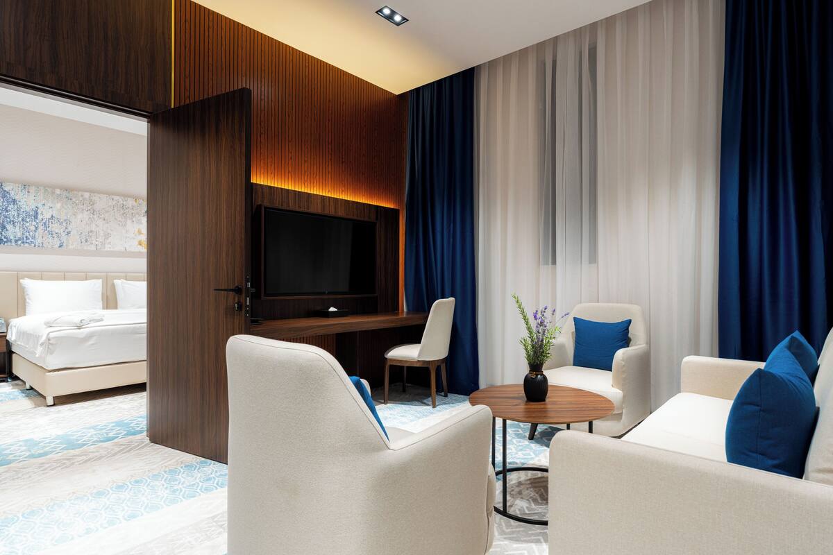 Una habitación de hotel moderna con una cama grande, televisión, zona de estar con sofás y sillones, y un ventanal con cortinas.