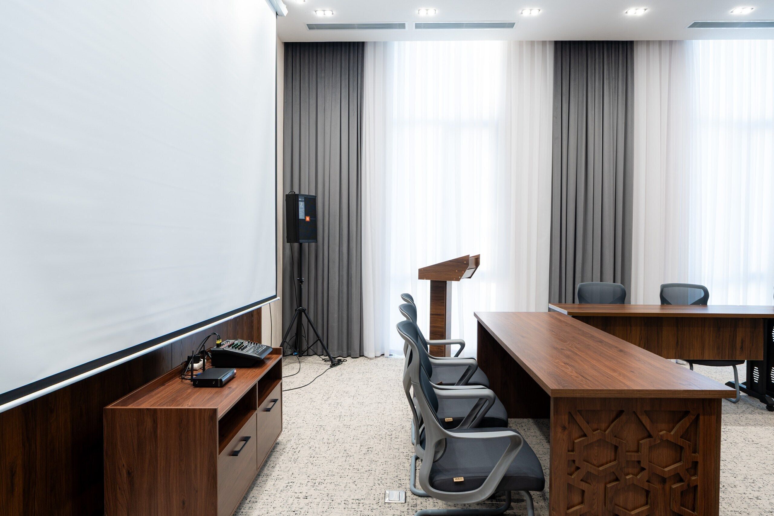 Une salle de conférence équipée d’un bureau en bois, de chaises, d’un écran de projection et d’un pupitre.