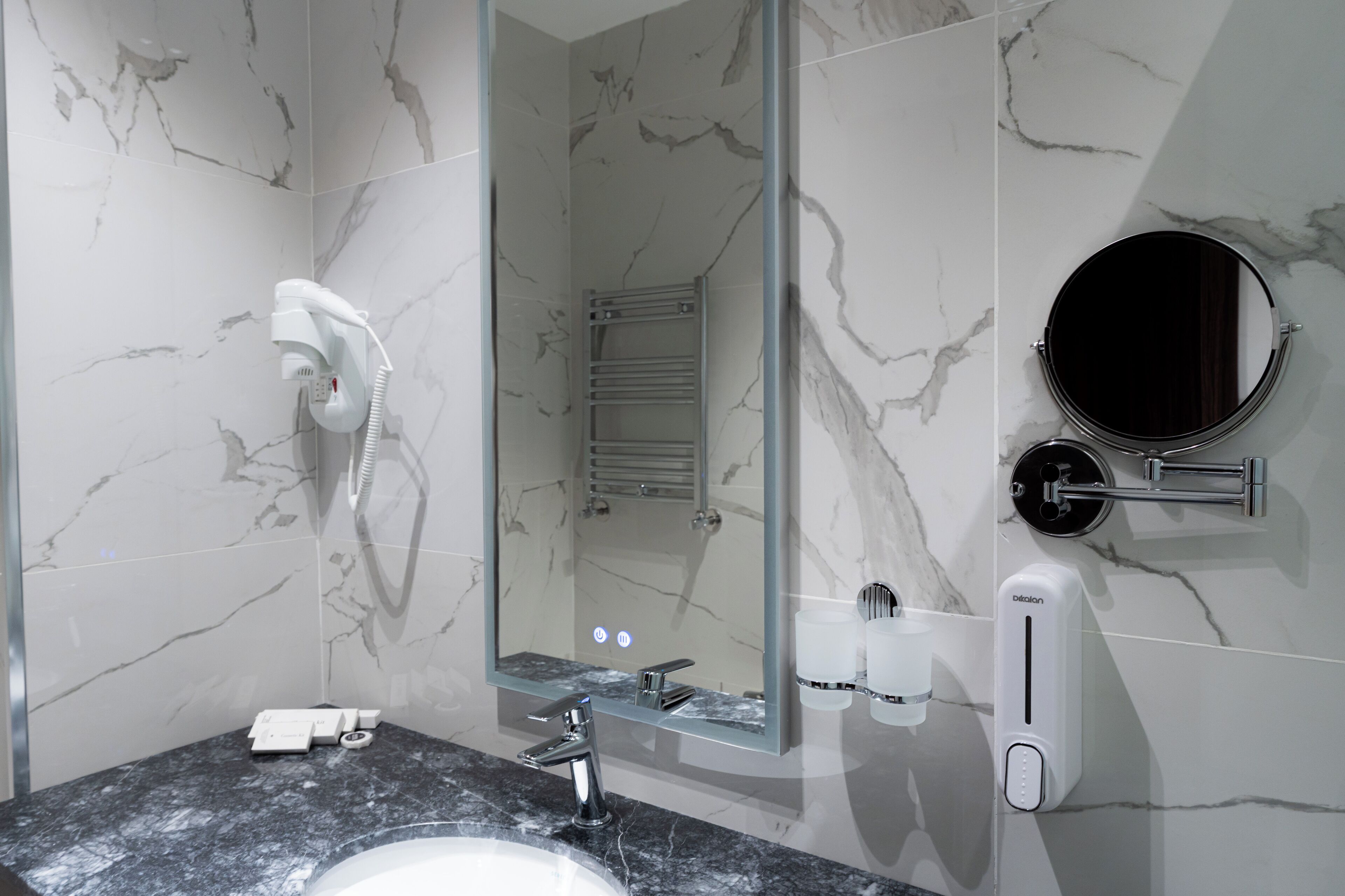 A modern bathroom with a marble backsplash, a built-in shower, a round mirror, a handheld showerhead, a towel rack, and a soap dispenser.