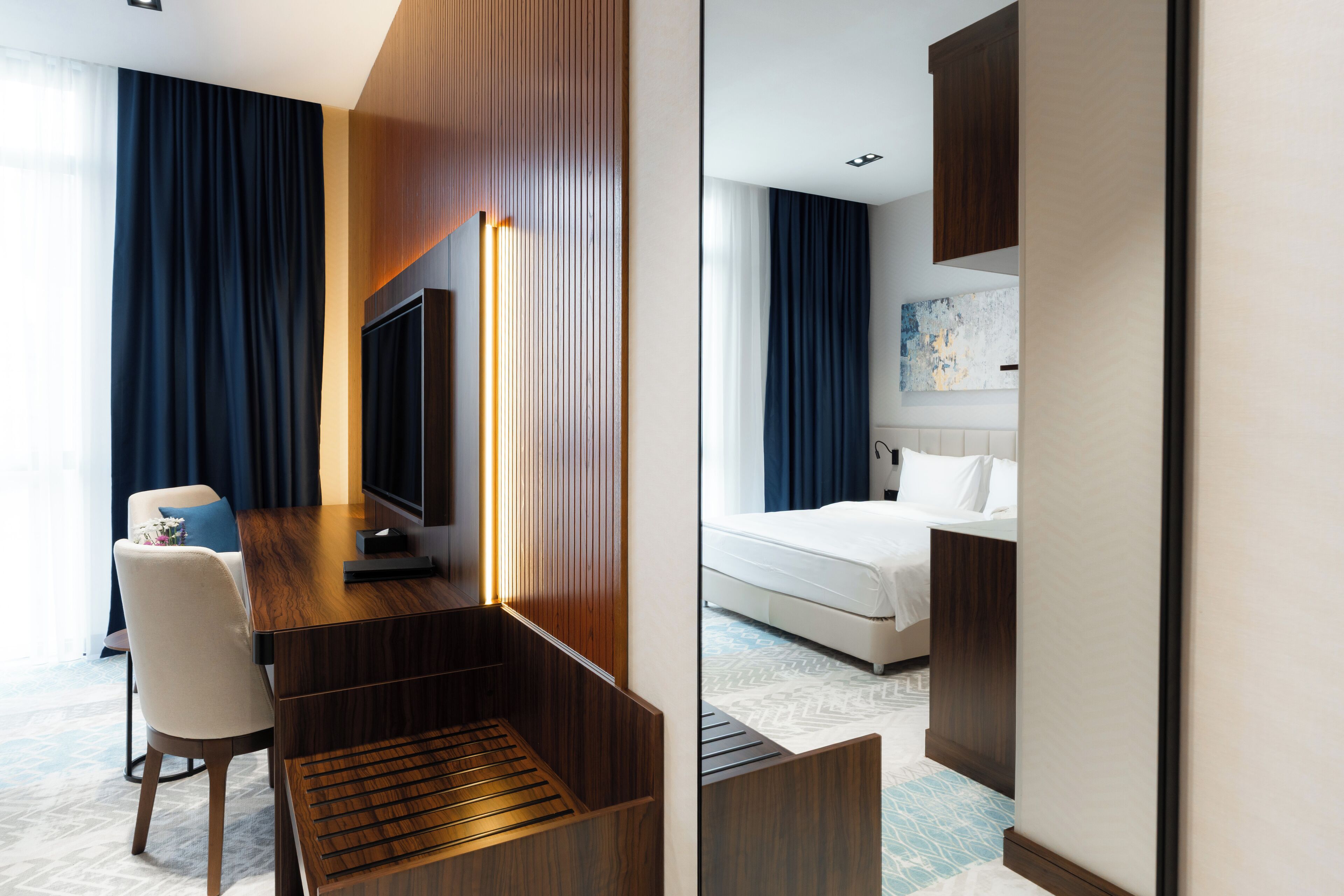 A hotel room with a wooden desk, a chair, a bed, and a large window with curtains.