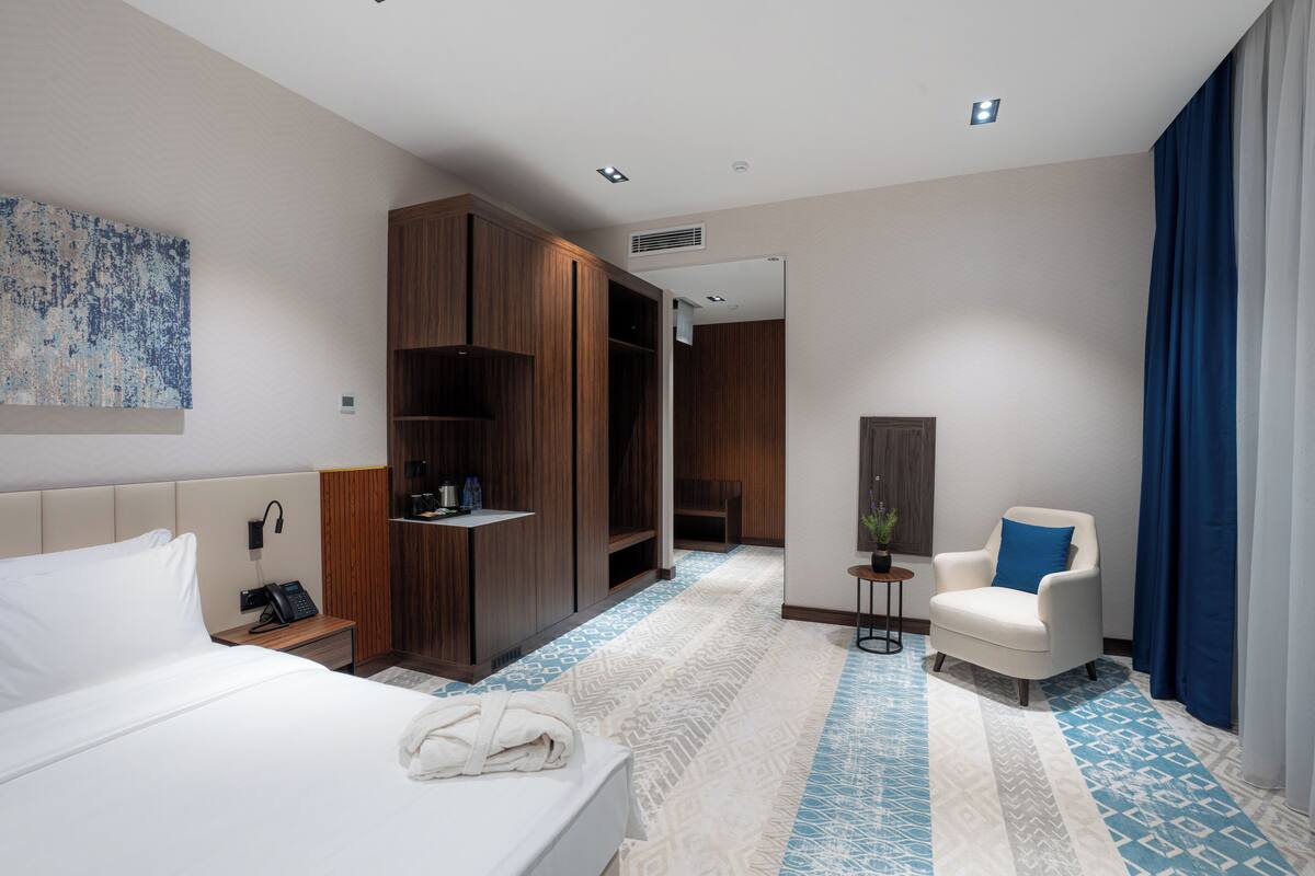 Habitación de hotel con una cama grande, un armario, zona de estar con una silla y una mesita con una planta.