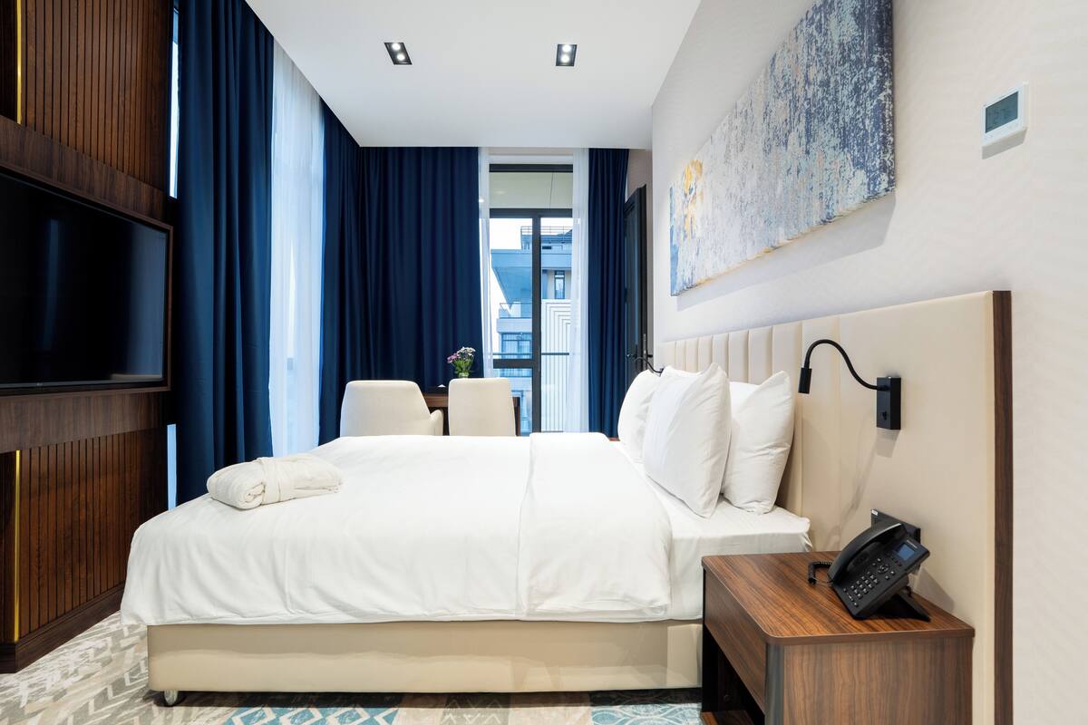 Habitación de hotel con una cama grande, un televisor de pantalla plana, teléfono junto a la cama y vista a la ciudad desde la ventana.