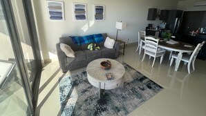 Een moderne woonkamer met een grijze bank, een ronde salontafel en een eethoek met witte stoelen. Er hangen drie abstracte schilderijen aan de muur en er is een plafondventilator.