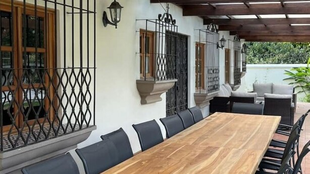 A covered outdoor dining area with a long wooden table and black chairs, surrounded by a pergola with wooden beams and metal railings. - Spacious oasis with pool and garden, minutes from Antigua. (Antigua Guatemala)