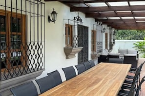 A covered outdoor dining area with a long wooden table and black chairs, surrounded by a pergola with wooden beams and metal railings.