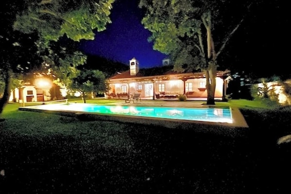 A house with a swimming pool at night, surrounded by trees and illuminated by exterior lights.