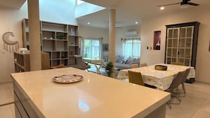 Un vaste espace de vie comprenant un îlot central, une table à manger avec des chaises, une bibliothèque et une grande fenêtre laissant entrer la lumière naturelle.