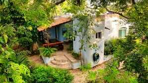 Un petit bâtiment blanc, doté d’un toit recouvert de tuiles, entouré d’une végétation luxuriante et accessible par un escalier menant à une porte.
