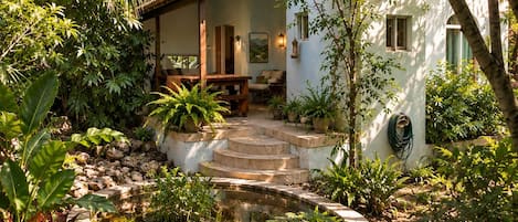A house with a stone pathway leading to a wooden deck, surrounded by lush greenery and a small pond.