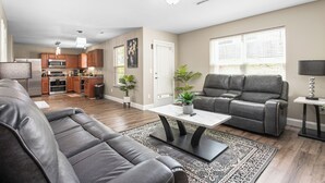 A modern living room with a gray leather sofa, a gray recliner, and a coffee table. There is a kitchen in the background with wooden cabinets and a stainless steel refrigerator. - 327 Beautiful Residence in Mebane NC. (Mebane)