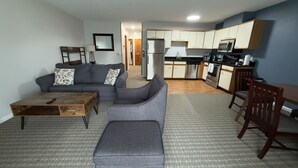 A compact living space with a sofa, ottoman, coffee table, and a dining area with a chair. A kitchen with appliances and cabinets is visible in the background. - Extended Stay- Full Kitchen 1BR apartment in Downtown Pittsfield (Pittsfield)