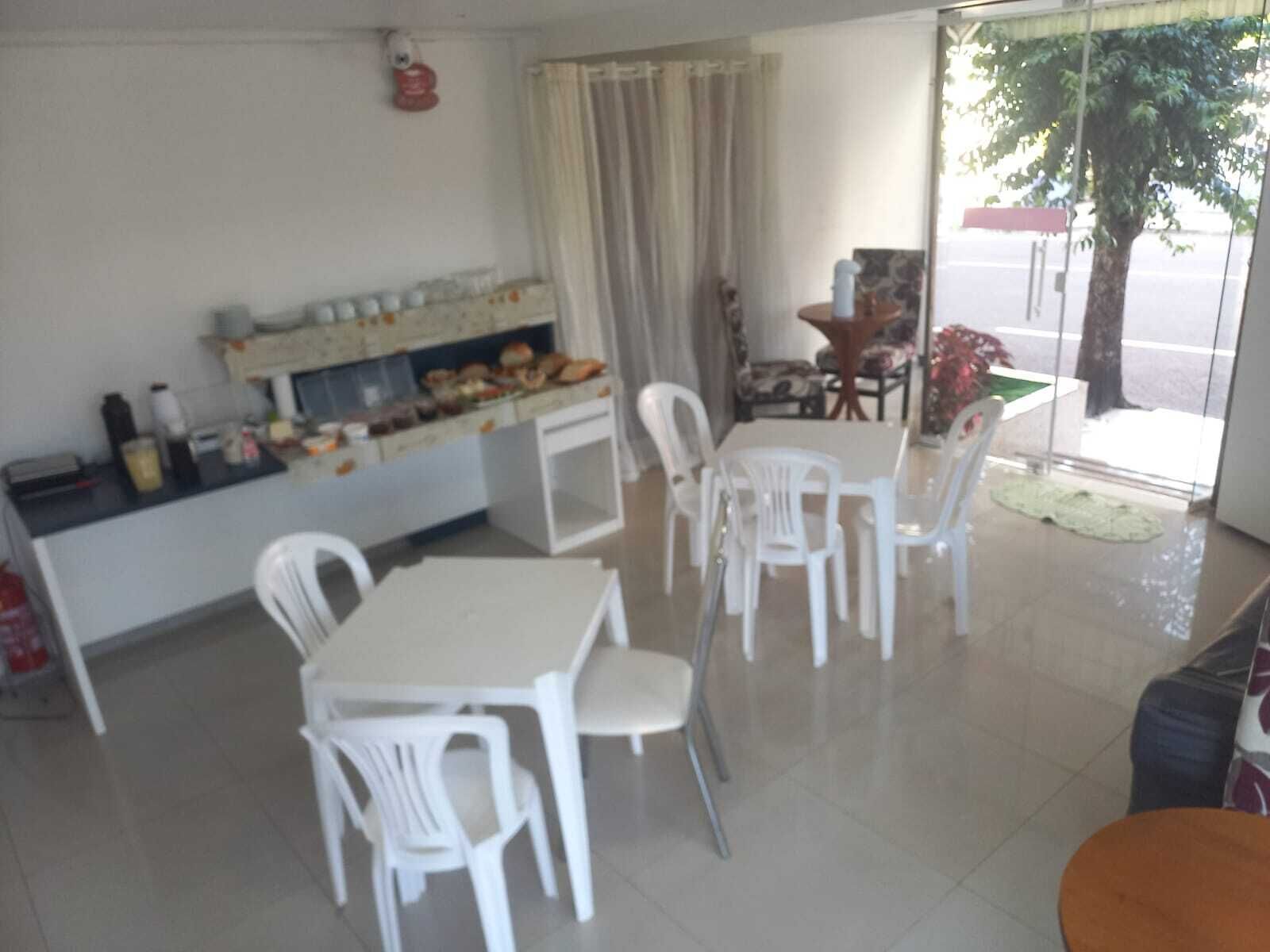 Un coin repas avec une table et des chaises blanches, un petit espace cuisine équipé d’un réfrigérateur et d’étagères, et une vue sur la rue.