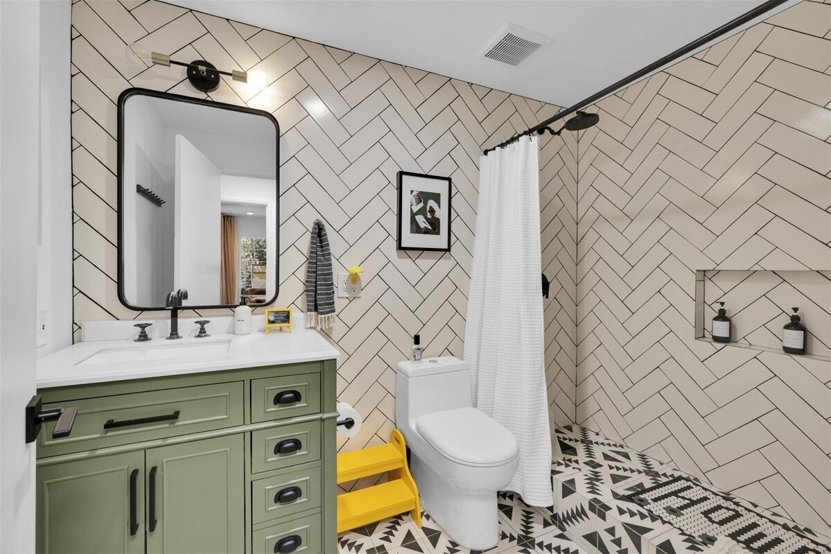 Une salle de bain avec un mur orné d’un motif en chevrons, un meuble-lavabo vert avec des tiroirs, des toilettes blanches et un rideau de douche.