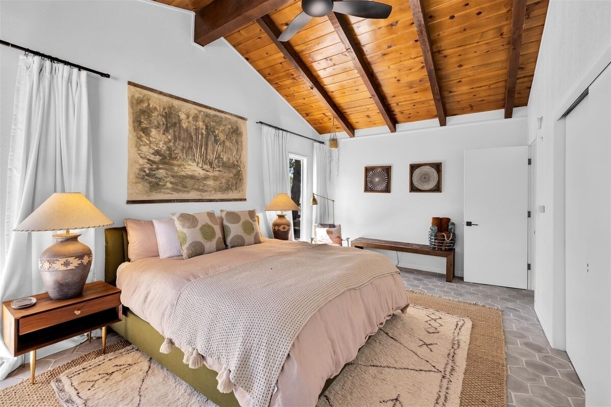 Ein Schlafzimmer mit Holzboden, einem großen Bett mit beiger Bettdecke, zwei Nachttischen mit Lampen, einem großen Gemälde an der Wand und einer Tür mit einem kleinen dekorativen Gegenstand.