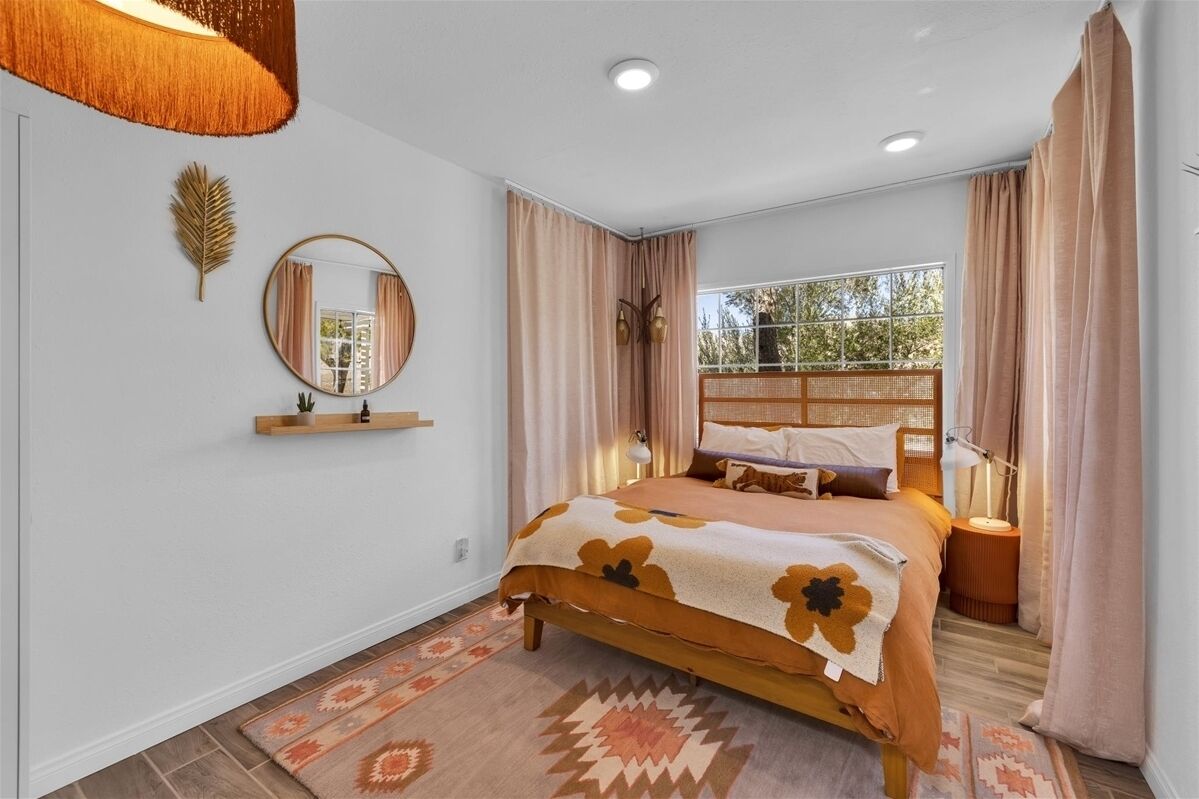 A bedroom with a bed, a window with curtains, a bedside lamp, and a wall-mounted mirror.