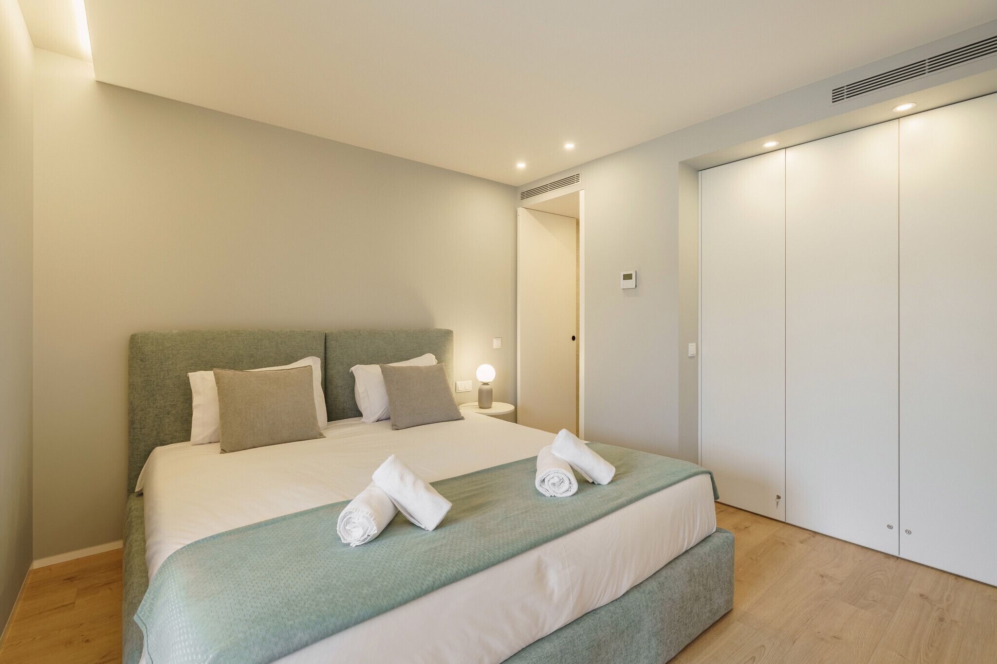 Una cama bien hecha con cabecera verde, ropa de cama blanca y dos toallas enrolladas sobre la cama.