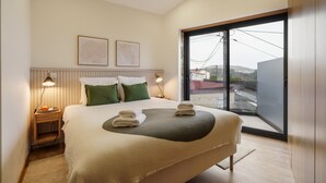 Een moderne slaapkamer met een groot bed, twee bedlampjes, uitzicht door een groot raam en twee ingelijste foto’s aan de muur.