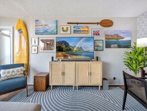 A living room with a large flat-screen TV, a surfboard, and various framed pictures on the wall. - Waikiki Penthouse 2bd/1ba, Diamond Head Views, 5 mins from Beach and Dining (Honolulu)