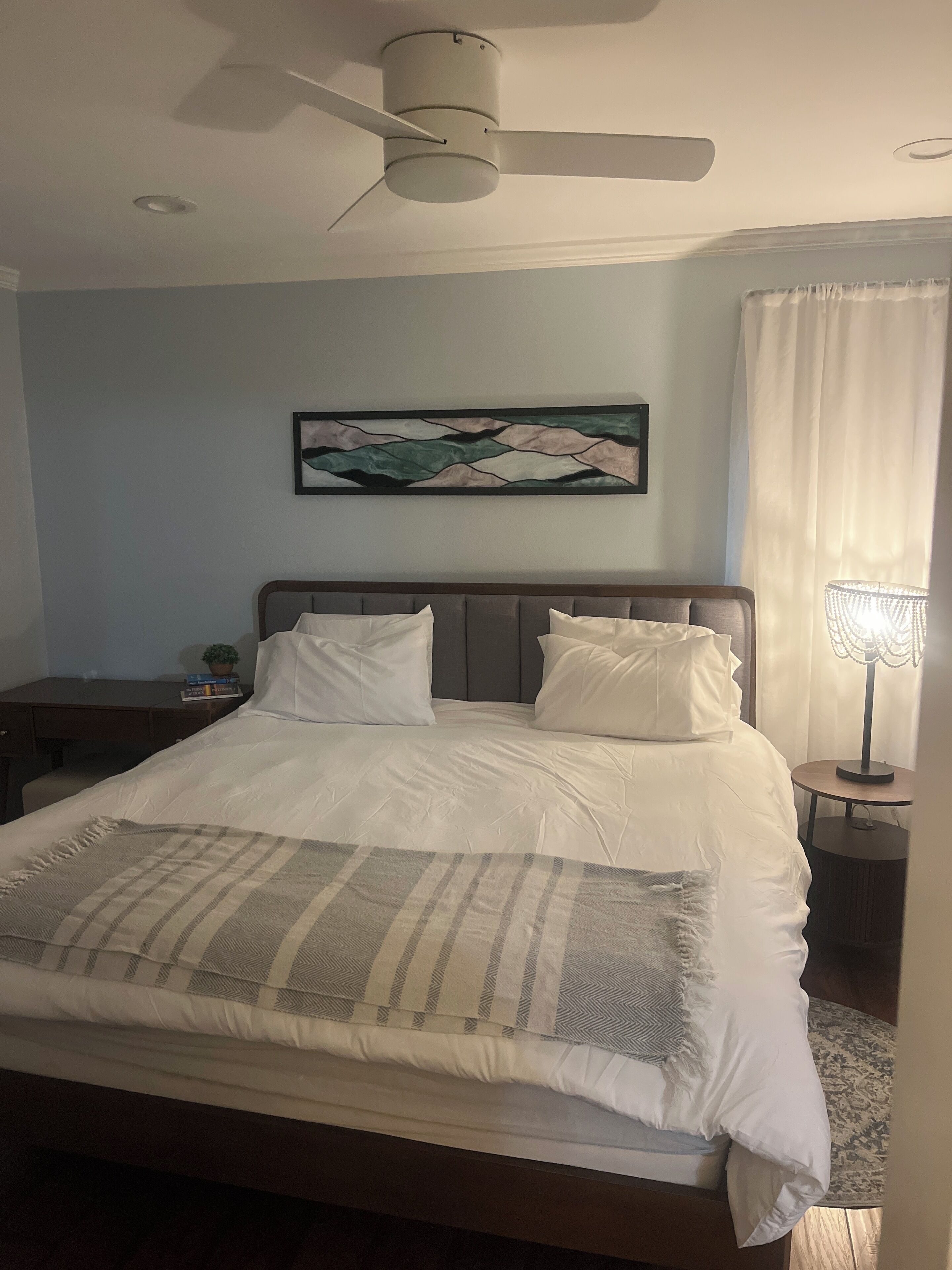 A bedroom with a bed, a ceiling fan, a nightstand, a lamp, and a painting on the wall.