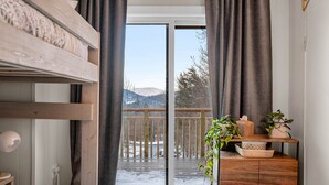 Ein Schlafzimmer mit Etagenbett, Nachttisch mit Lampe, Kommode und Blick auf eine verschneite Terrasse und Berge.