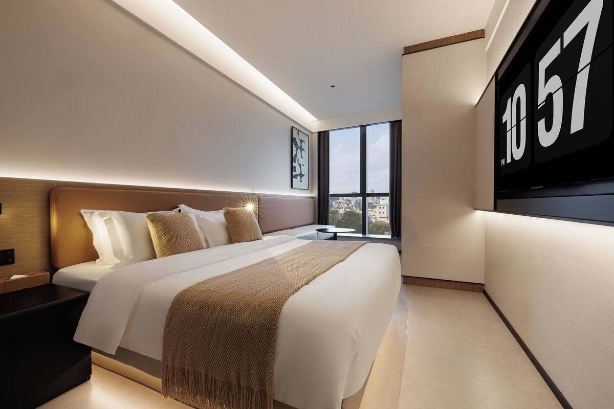 Une chambre d’hôtel moderne avec un grand lit, une table de chevet, une petite table ronde et une grande fenêtre donnant sur la ville.