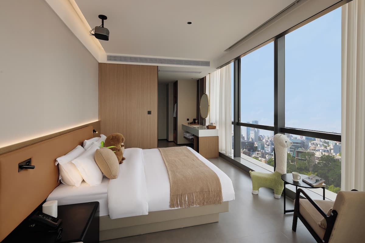 Une chambre d’hôtel moderne avec un grand lit, une table de chevet, un petit bureau, une chaise, une grande fenêtre donnant sur la ville et un miroir.