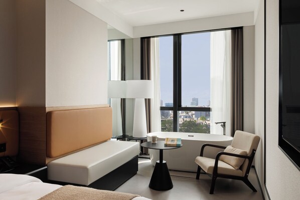 Une chambre d’hôtel moderne, dotée d’un canapé, d’un fauteuil, d’une petite table et d’une grande fenêtre donnant sur la ville.