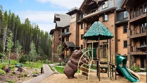 Ein Kinderspielplatz mit Rutsche und Klettergerüsten, umgeben von Wald und einem mehrstöckigen Gebäude mit Balkonen.