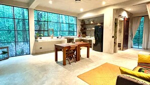 A spacious living area with a large window overlooking a forest, a dining table, and a kitchen area. - Beautiful House in the Heart of the Jungle - Yaáxnik (Tulum)