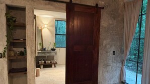 A bedroom with a large bed, a wooden door, and a view of a forest through a window. - Beautiful House in the Heart of the Jungle - Yaáxnik (Tulum)